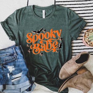 May include: A green t-shirt with the text "Spooky Babe" in orange lettering. The shirt has a distressed look and is styled with denim shorts and brown boots.
