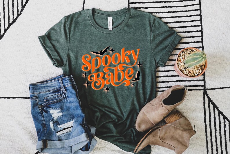 May include: A green t-shirt with the text "Spooky Babe" in orange lettering. The shirt has a distressed look and is styled with denim shorts and brown boots.