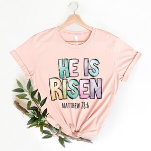 May include: A pink t-shirt with the words "He is Risen" in colorful, watercolor-style letters. The text "Matthew 28:6" is printed below the main text.