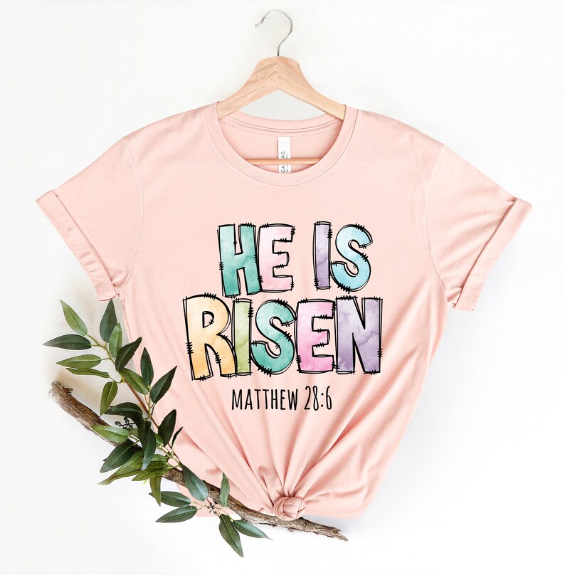 May include: A pink t-shirt with the words "He is Risen" in colorful, watercolor-style letters. The text "Matthew 28:6" is printed below the main text.
