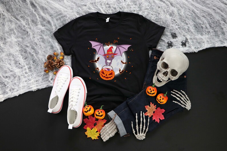 May include: Black t-shirt with a cartoon bat wearing a witch hat and holding a pumpkin. The bat is flying in front of a full moon. There are small bats flying around the moon.