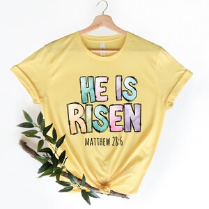 May include: Yellow t-shirt with a colorful graphic that says "He is Risen" with the scripture reference "Matthew 28:6" underneath.