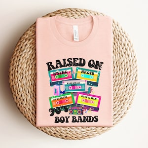 May include: Pink t-shirt with the text "RAISED ON 90's BOY BANDS" and a graphic of cassette tapes with the names of boy bands from the 1990s, including N'SYNC, NKOTB, Backstreet Boys, 38 Degrees, and Boyz II Men.