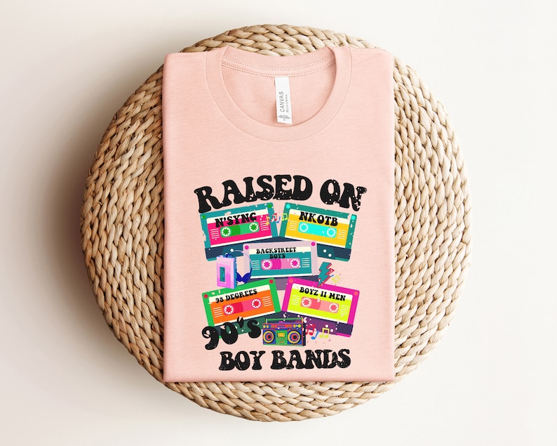 May include: Pink t-shirt with the text "RAISED ON 90's BOY BANDS" and a graphic of cassette tapes with the names of boy bands from the 1990s, including N'SYNC, NKOTB, Backstreet Boys, 38 Degrees, and Boyz II Men.