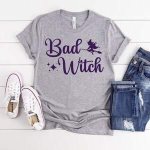 Puede incluir: Una camiseta gris con el texto "Bad Witch" en letra morada. Una silueta de bruja vuela en una escoba sobre el texto. La camiseta est&aacute; colocada sobre una superficie blanca con un par de zapatillas blancas y unos vaqueros azules.
