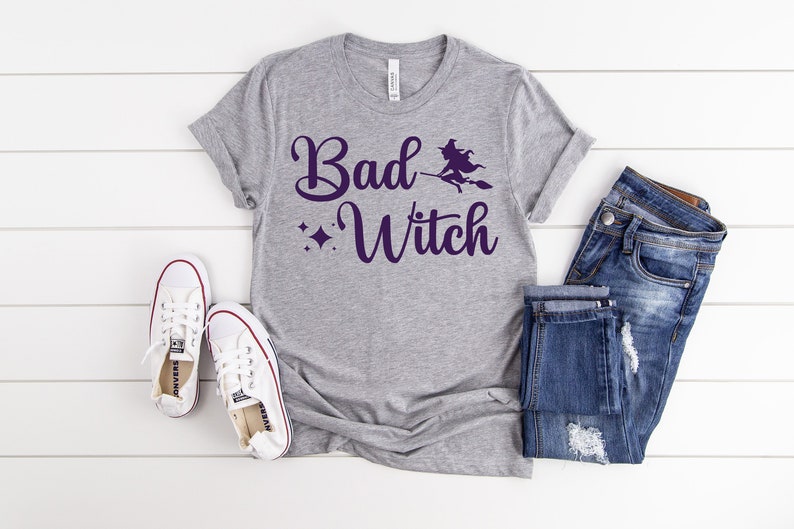 Puede incluir: Una camiseta gris con el texto "Bad Witch" en letra morada. Una silueta de bruja vuela en una escoba sobre el texto. La camiseta est&aacute; colocada sobre una superficie blanca con un par de zapatillas blancas y unos vaqueros azules.