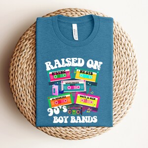 May include: A teal blue t-shirt with the text "Raised on 90's Boy Bands" and a graphic of cassette tapes with the names of popular boy bands from the 1990s, including NSYNC, NKOTB, Backstreet Boys, 3 Degrees, and Boyz II Men.