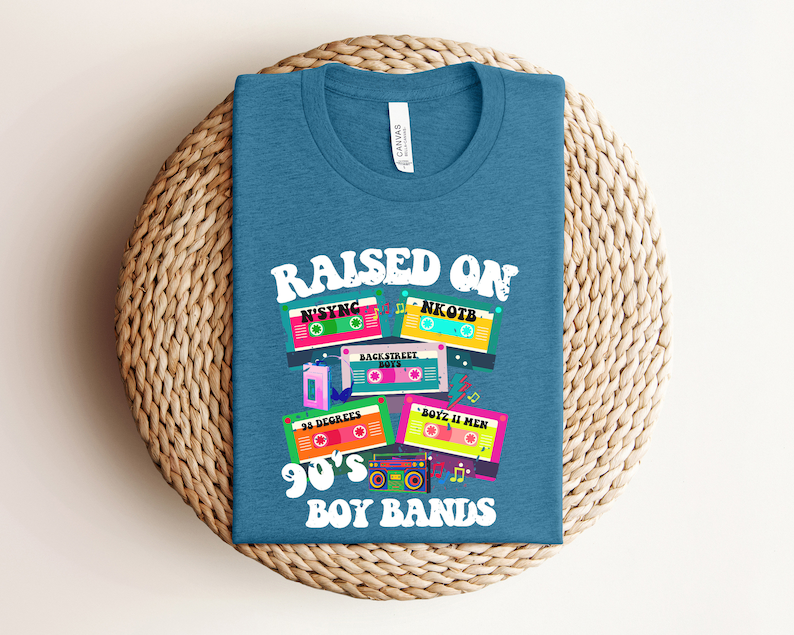 May include: A teal blue t-shirt with the text "Raised on 90's Boy Bands" and a graphic of cassette tapes with the names of popular boy bands from the 1990s, including NSYNC, NKOTB, Backstreet Boys, 3 Degrees, and Boyz II Men.