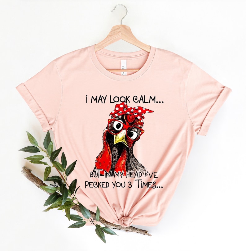 May include: A pink t-shirt with a graphic of a red and black chicken wearing a red bandana with white polka dots and red glasses. The text on the shirt reads "I may look calm... But in my head I've pecked you 3 times..."