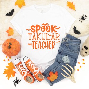 May include: White t-shirt with the text "SPOOK TAKULAR TEACHER" in orange lettering. The shirt is surrounded by Halloween decorations, including a pumpkin, leaves, spiders, and a skull.