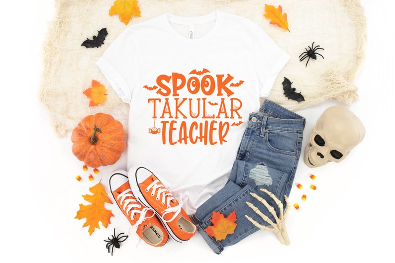 May include: White t-shirt with the text "SPOOK TAKULAR TEACHER" in orange lettering. The shirt is surrounded by Halloween decorations, including a pumpkin, leaves, spiders, and a skull.
