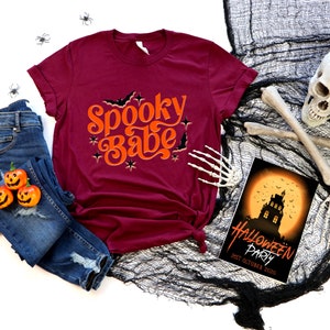 May include: A maroon t-shirt with the text "Spooky Babe" in orange lettering with bats and stars. The shirt is laid out on a white background with a pair of blue jeans, three orange jack-o-lantern pumpkins, and a black spiderweb with a skull and crossbones.