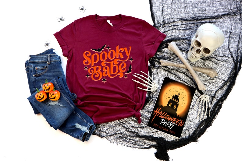 May include: A maroon t-shirt with the text "Spooky Babe" in orange lettering with bats and stars. The shirt is laid out on a white background with a pair of blue jeans, three orange jack-o-lantern pumpkins, and a black spiderweb with a skull and crossbones.