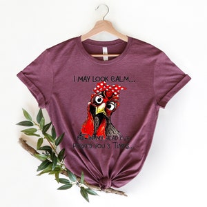 May include: A maroon t-shirt with a graphic of a chicken wearing glasses and a red bandana with white polka dots. The text on the shirt reads "I may look calm... But in my head I've pecked you 3 times..."