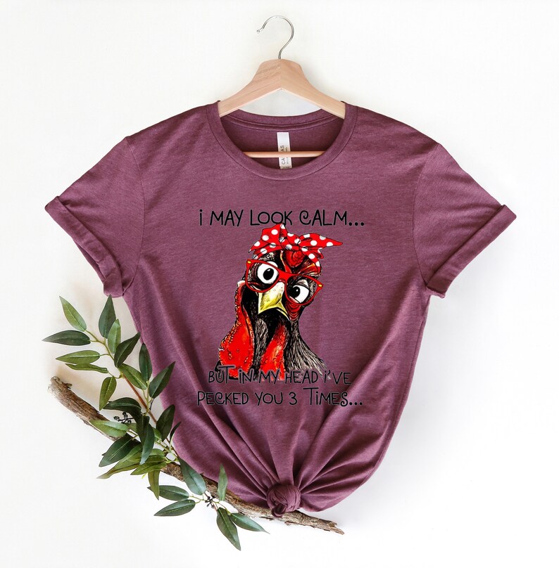 May include: A maroon t-shirt with a graphic of a chicken wearing glasses and a red bandana with white polka dots. The text on the shirt reads "I may look calm... But in my head I've pecked you 3 times..."