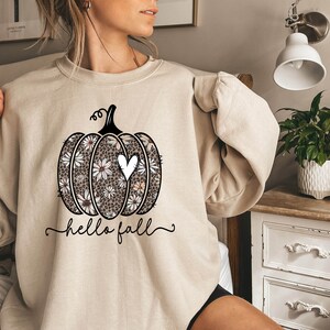 Puede incluir: Una sudadera beige con un gr&aacute;fico de una calabaza con un coraz&oacute;n blanco y un patr&oacute;n floral. El texto "hello fall" est&aacute; impreso debajo de la calabaza.
