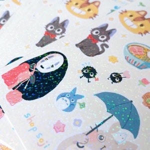 May include: Sheets of glittery stickers featuring various cartoon characters. Designs include a black cat with a red bow, a character with a white mask, and a blue umbrella. The text "shop soji" is visible.