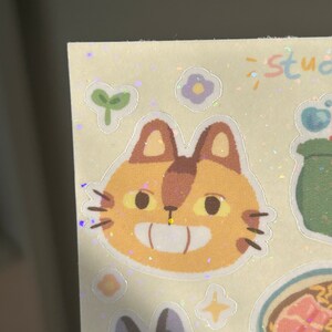 May include: A sheet of stickers featuring cartoon illustrations. The stickers include a smiling cat face with brown ears and whiskers, a black cat, a bowl of ramen, and other whimsical designs. The word "studio" is written in pastel colors.