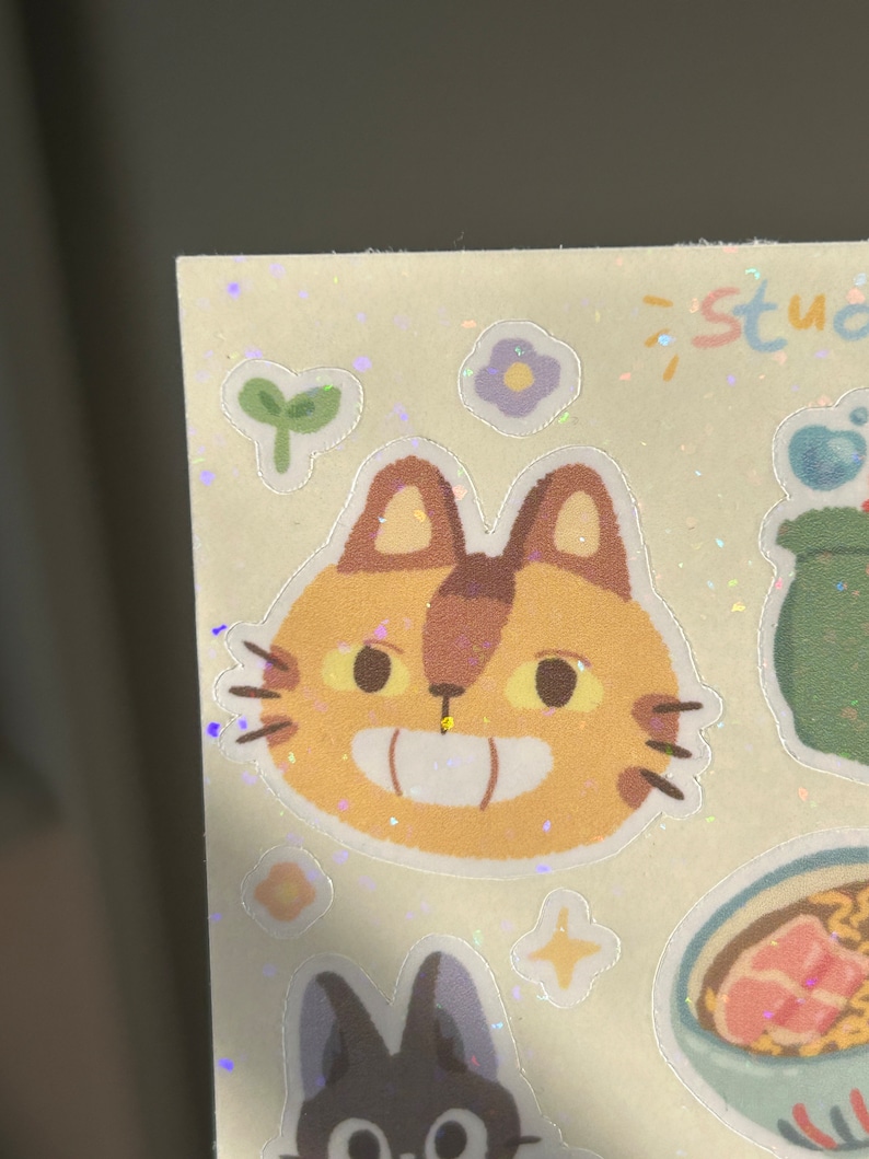May include: A sheet of stickers featuring cartoon illustrations. The stickers include a smiling cat face with brown ears and whiskers, a black cat, a bowl of ramen, and other whimsical designs. The word "studio" is written in pastel colors.