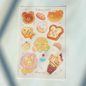 May include: A sticker sheet featuring various baked goods with animal faces. Designs include a bear-shaped bun, a fish-shaped pastry, a turtle-shaped pastry, and a lion-shaped pastry. The text "bakery eats" and "shop soji" are also visible.