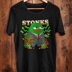 May include: Black t-shirt featuring a cartoon frog in a suit holding a briefcase, surrounded by money and arrows. The word "STONKS" is displayed above the frog in yellow and green. The shirt is hanging on a wooden hanger.