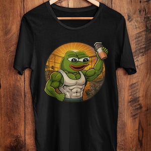 Könnte beinhalten: Schwarzes T-Shirt mit einem Cartoon-Frosch, der einen weißen Tanktop trägt und einen Shaker hält. Der Frosch ist muskulös und vor einem kreisförmigen Hintergrund mit einem Fitnessstudio abgebildet. Das T-Shirt hängt an einem Holzbügel.