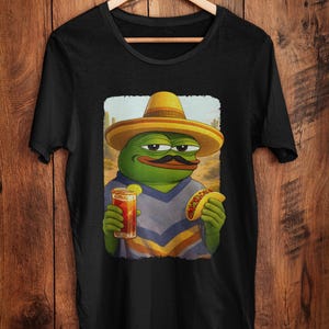 May include: Black t-shirt with a cartoon frog wearing a sombrero and a moustache, holding a drink and a taco. The frog is set against a desert backdrop. The t-shirt is displayed on a wooden hanger.