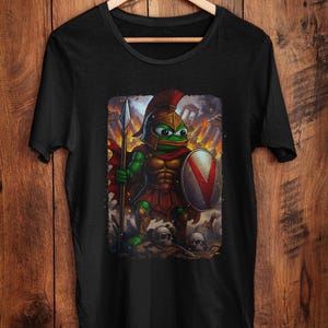 May include: Black t-shirt featuring a cartoon frog warrior in a Roman-style helmet and armor, holding a spear and shield. The frog is green and gold, with a red cape. The background shows a battle scene with skulls.