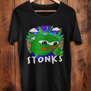 May include: Black t-shirt with a green cartoon frog and the word "STONKS" in white. The frog is surrounded by dollar signs and money, set against a blue and purple backdrop.