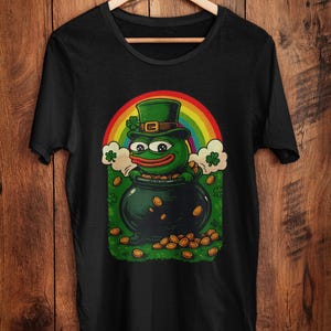 May include: Black t-shirt featuring a cartoon frog wearing a green leprechaun hat, emerging from a pot of gold coins. A rainbow arches above the pot, with shamrocks and clouds on either side. The design is centered on the shirt.