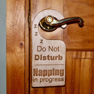 May include: A wooden door hanger with the text "Do Not Disturb Napping in progress" and three Z's above the text. The hanger is attached to a wooden door with a gold door handle.
