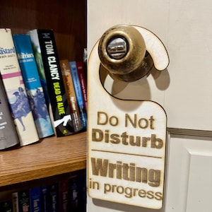 Puede incluir: Un colgador de puerta de madera con el texto "No molestar Escritura en curso" en letras marrones. El colgador está colgado en una puerta blanca con un pomo de puerta dorado.