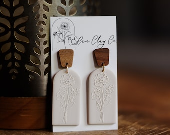 May include: A pair of cream-colored, floral-embossed earrings with wooden tops. The earrings are rectangular with an arched top and feature a delicate flower design. The earrings are connected with gold-colored hardware. The brand name "Eden Clay Co" is visible on the card.
