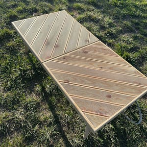 May include: A light wood folding table with a diagonal slat pattern. The table is made of two sections that fold together. The table is sitting on a grassy surface.