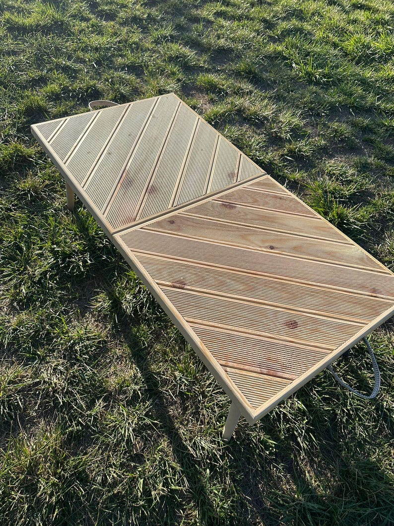 May include: A light wood folding table with a diagonal slat pattern. The table is made of two sections that fold together. The table is sitting on a grassy surface.