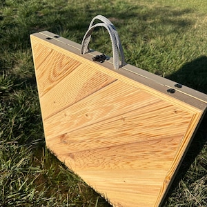 May include: A wooden folding table with a light brown finish and a gray fabric handle. The table is made of wood planks with a chevron pattern. The table is closed and folded up.