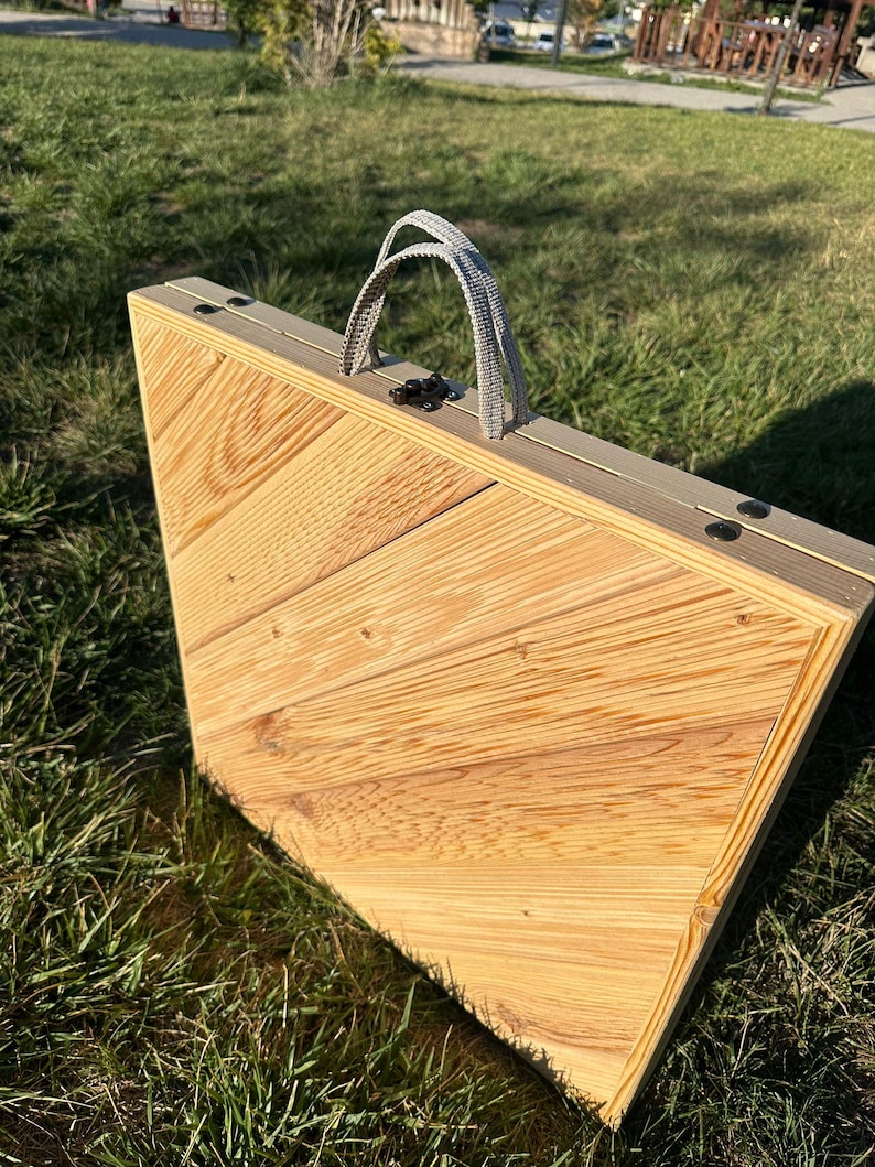 May include: A wooden folding table with a light brown finish and a gray fabric handle. The table is made of wood planks with a chevron pattern. The table is closed and folded up.