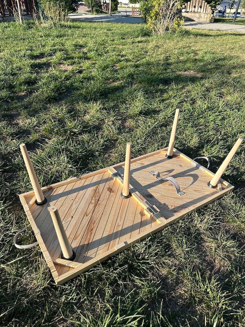 May include: A wooden folding table with a chevron pattern on the top. The table is folded up and has four legs that are attached to the underside of the table. The table is sitting on a grassy surface.