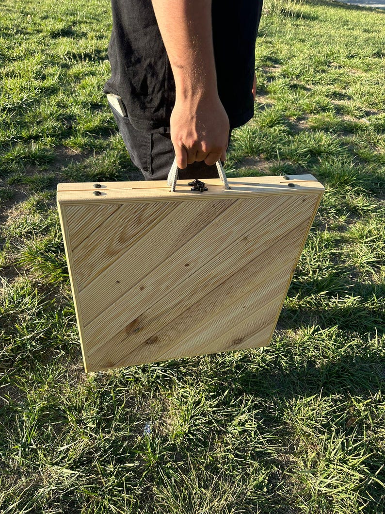 May include: A wooden folding table with a natural wood finish and a carrying handle. The table is closed and being held by a person's hand.