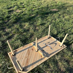 May include: A wooden folding table with a chevron pattern on the top. The table has four legs and a strap handle. The table is folded up and lying on the grass.