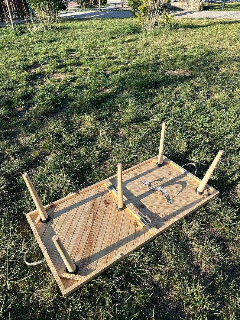 May include: A wooden folding table with a chevron pattern on the top. The table has four legs and a strap handle. The table is folded up and lying on the grass.