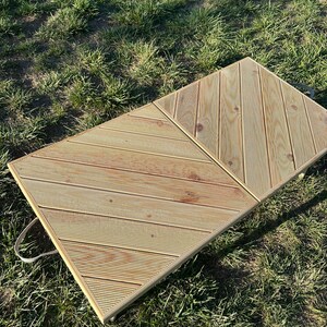 May include: A wooden folding table with a diagonal pattern on the top. The table is made of light brown wood and has two handles on the side. The table is sitting on a green grassy surface.