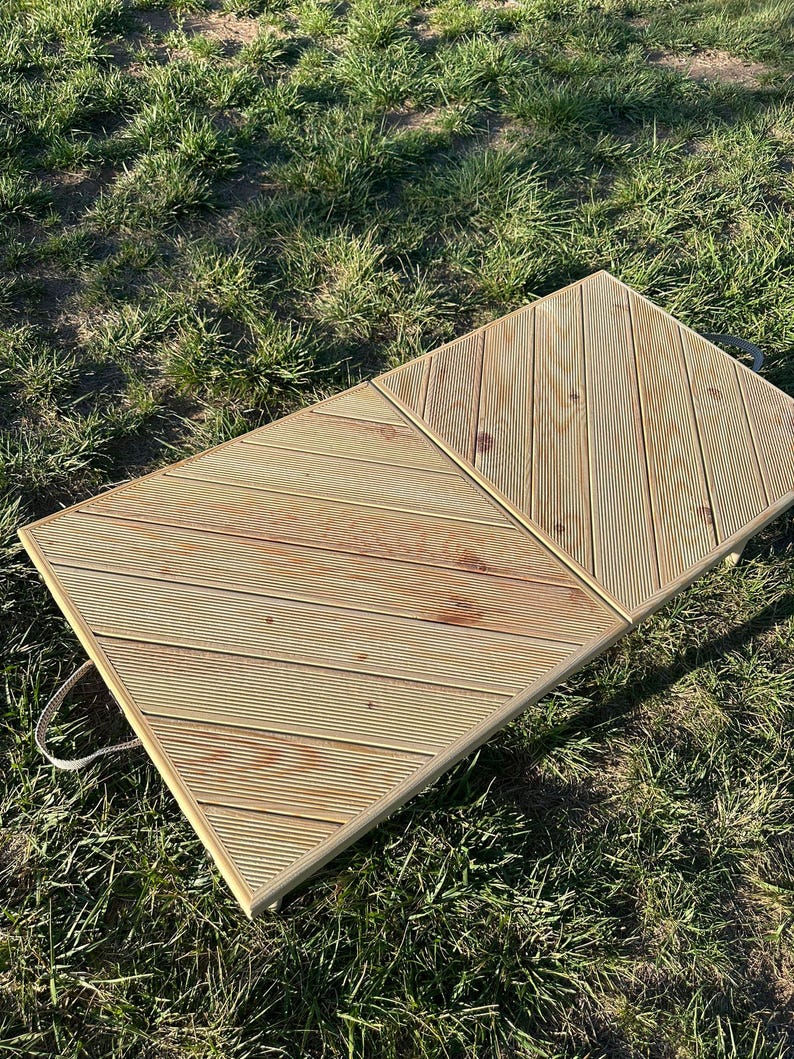 May include: A wooden folding table with a diagonal pattern on the top. The table is made of light brown wood and has two handles on the side. The table is sitting on a green grassy surface.