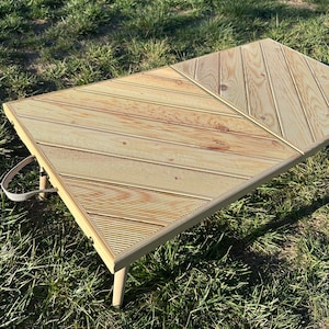 May include: A light brown wooden folding table with a diagonal pattern. The table has a white handle and is sitting on green grass.