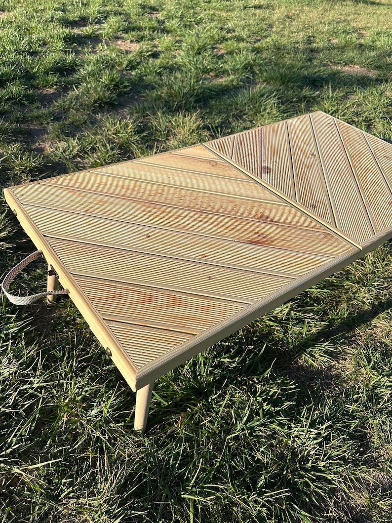 May include: A light brown wooden folding table with a diagonal pattern. The table has a white handle and is sitting on green grass.