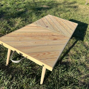 May include: A wooden folding table with a chevron pattern top and white painted legs. The table is sitting on a grassy surface.
