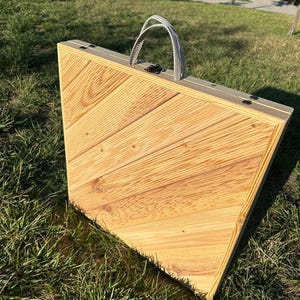May include: A wooden folding table with a light brown finish and a gray fabric handle. The table is closed and sitting on a grassy surface.
