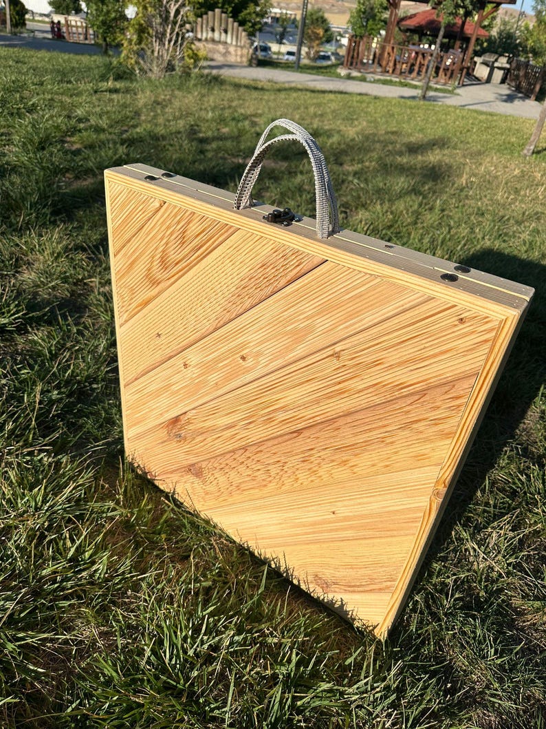 May include: A wooden folding table with a light brown finish and a gray fabric handle. The table is closed and sitting on a grassy surface.