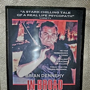May include: DVD cover art for the movie "In Broad Daylight" starring Brian Dennehy. The cover features a close-up of Brian Dennehy holding a gun, with the title of the movie in red letters and the tagline "A Terrifying True Story".