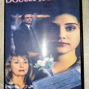 May include: DVD case cover for the movie "Double Jeopardy" featuring a woman with dark hair and a man in a suit. The woman is looking at the camera with a serious expression. The man is looking off to the side. The background is blurry and out of focus. The DVD case cover also features a tombstone with the text "Julia Mann Neilson" on it.